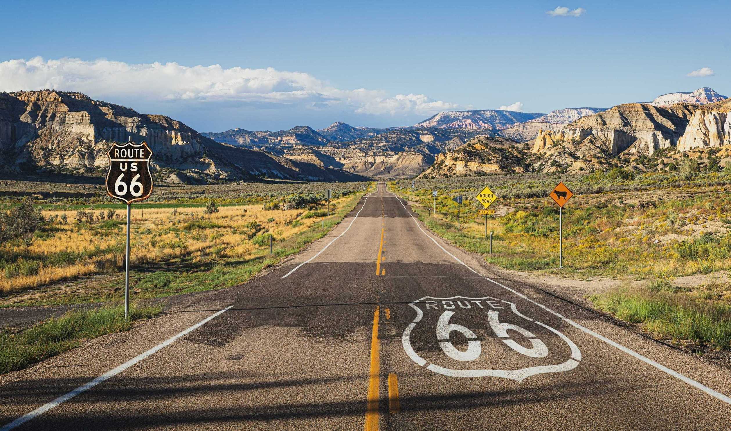 Scenic Route 66 highway stretching into the distance through a vast desert landscape with rocky mesas, golden grasslands under a blue sky.