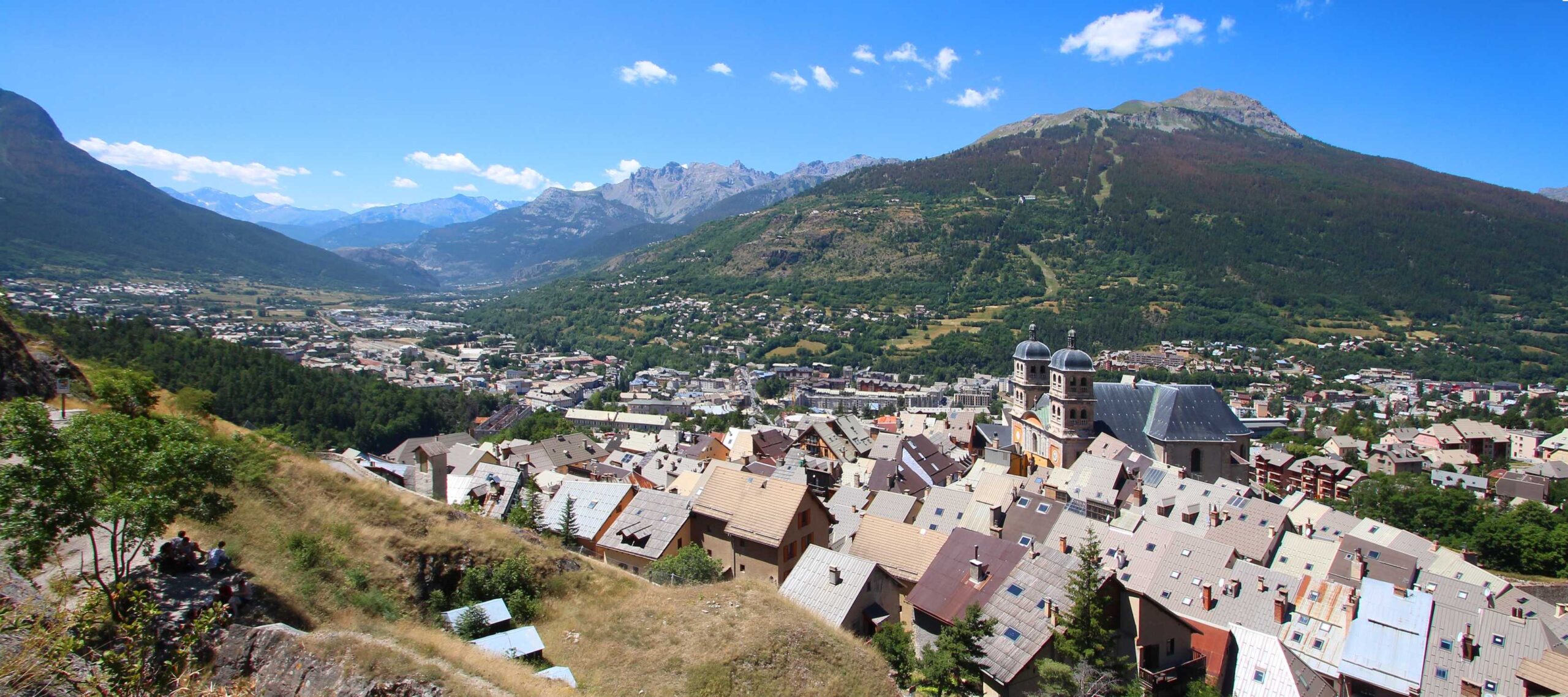 View of Briançon with clustered rooftops framed by lush green mountains under a blue sky.