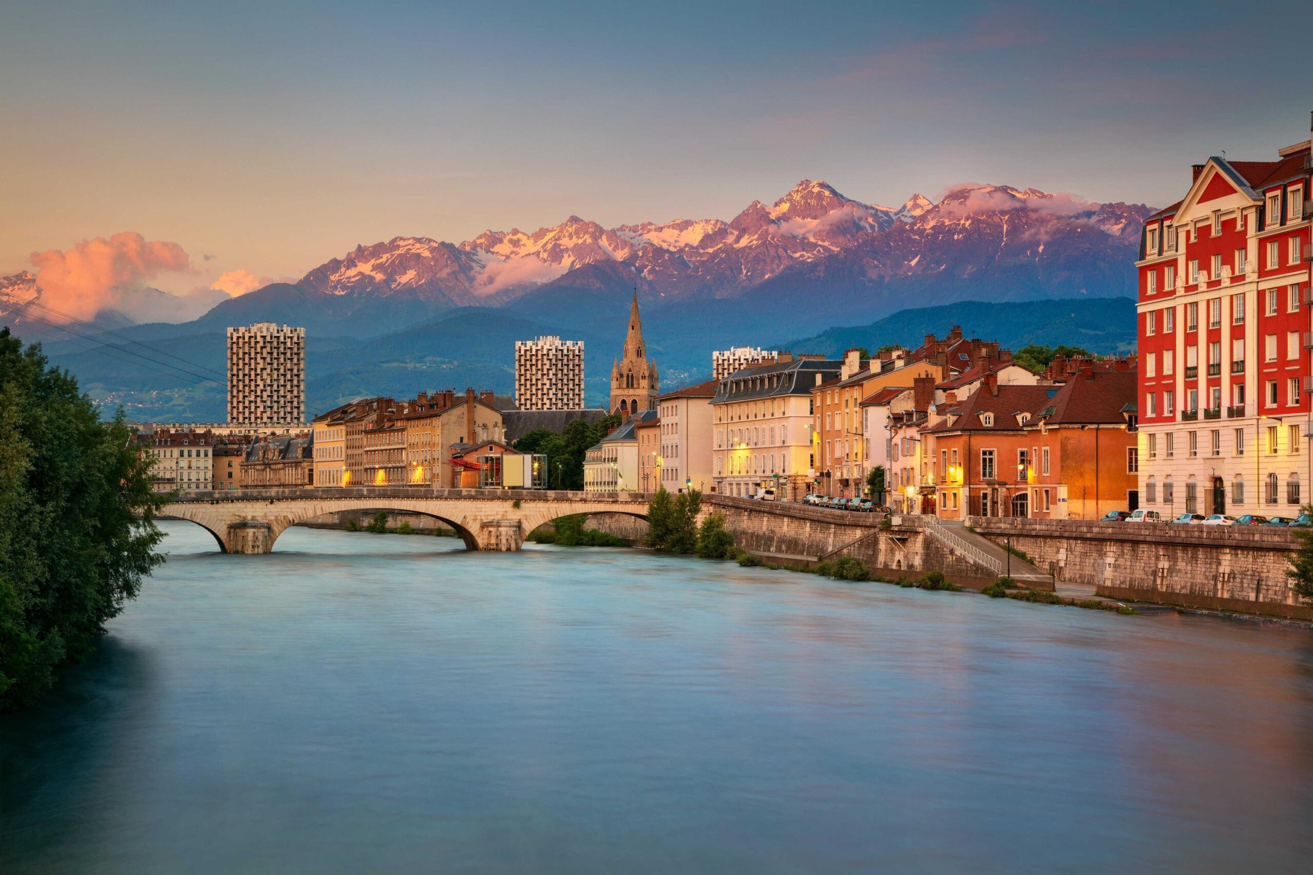 View of Briançon with clustered rooftops framed by lush green mountains under a blue sky.