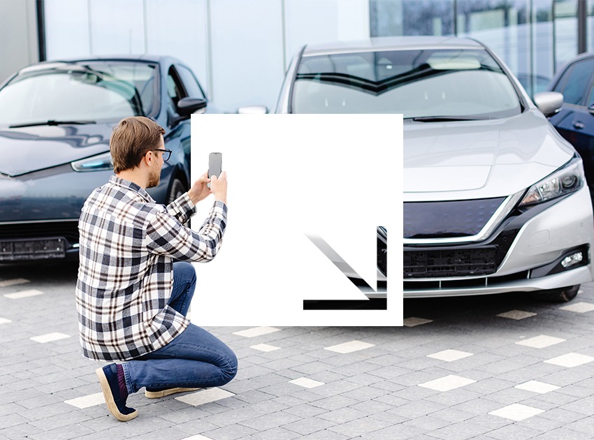 A man in a plaid shirt and jeans crouches while taking a photo of a silver car with his smartphone.